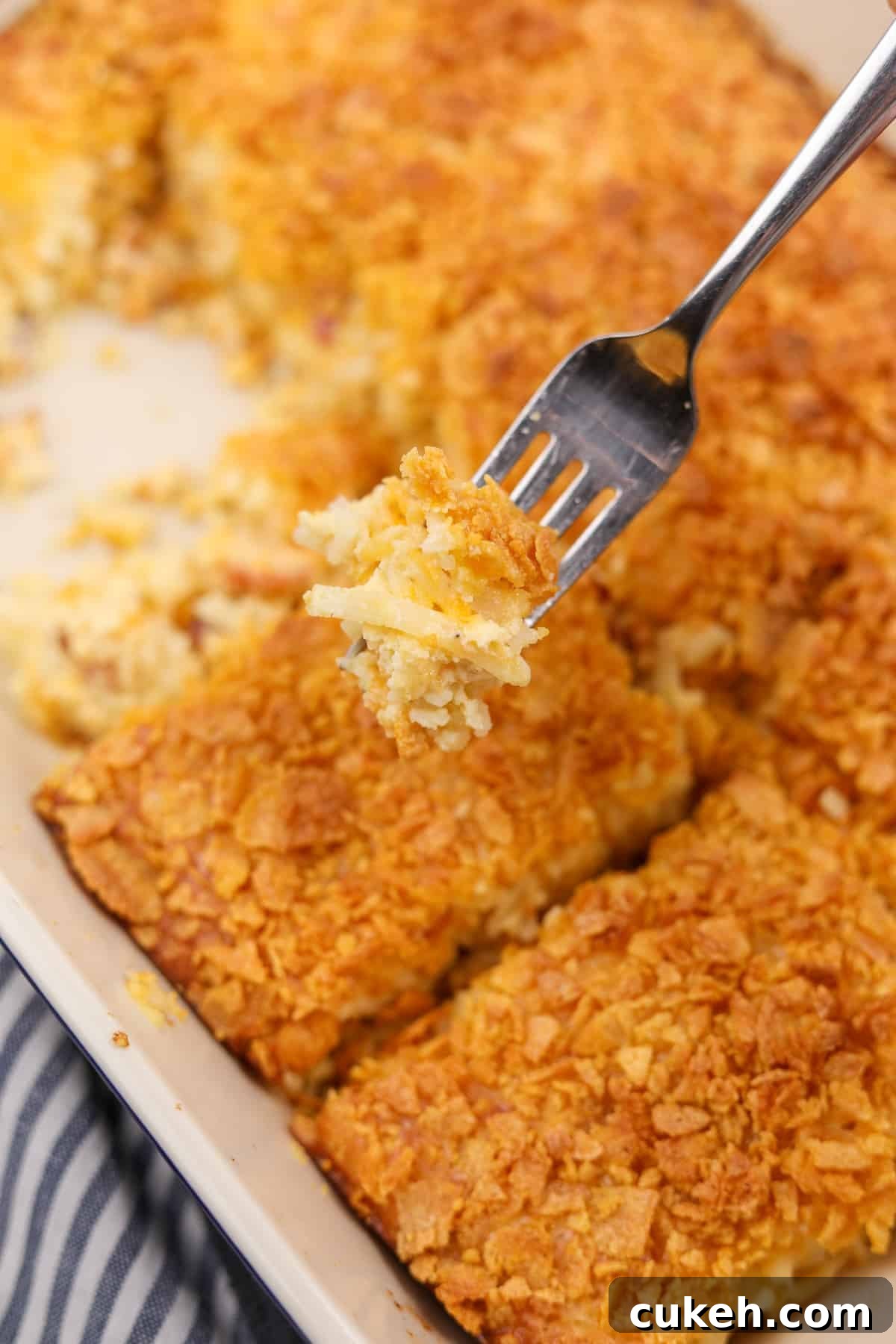A fork showing the texture of a shredded hashbrown casserole, which is in the background and topped with crunchy cornflakes.