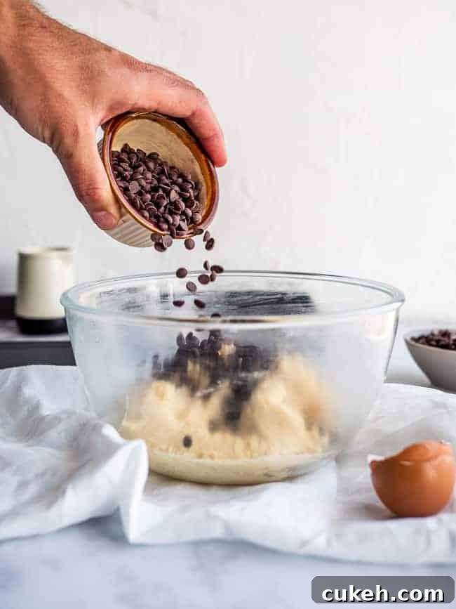 hand pouring chocolate chips into cookie dough inside a glass bowl.