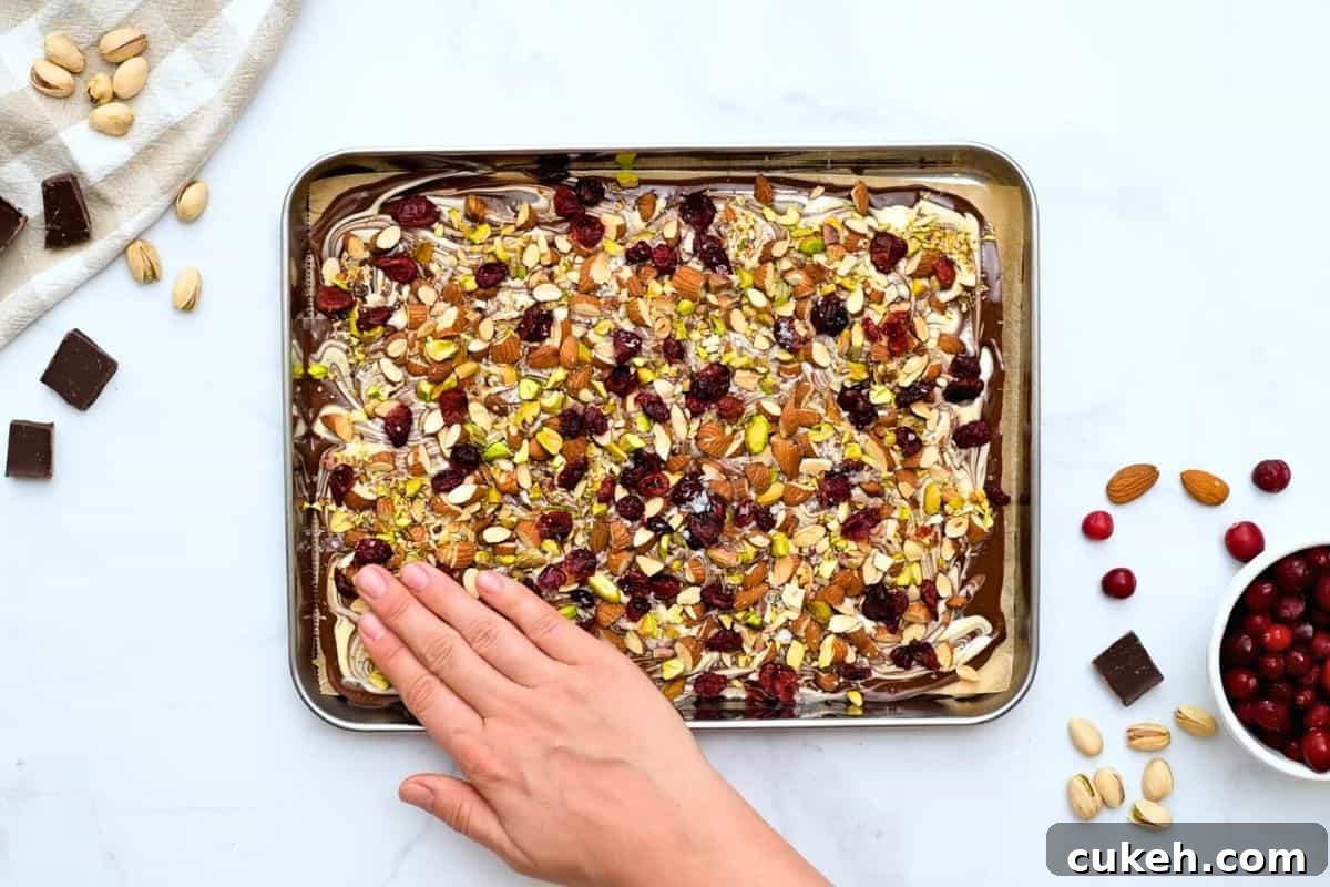 Hand gently pressing toasted nuts and dried cranberries onto marbled chocolate bark.