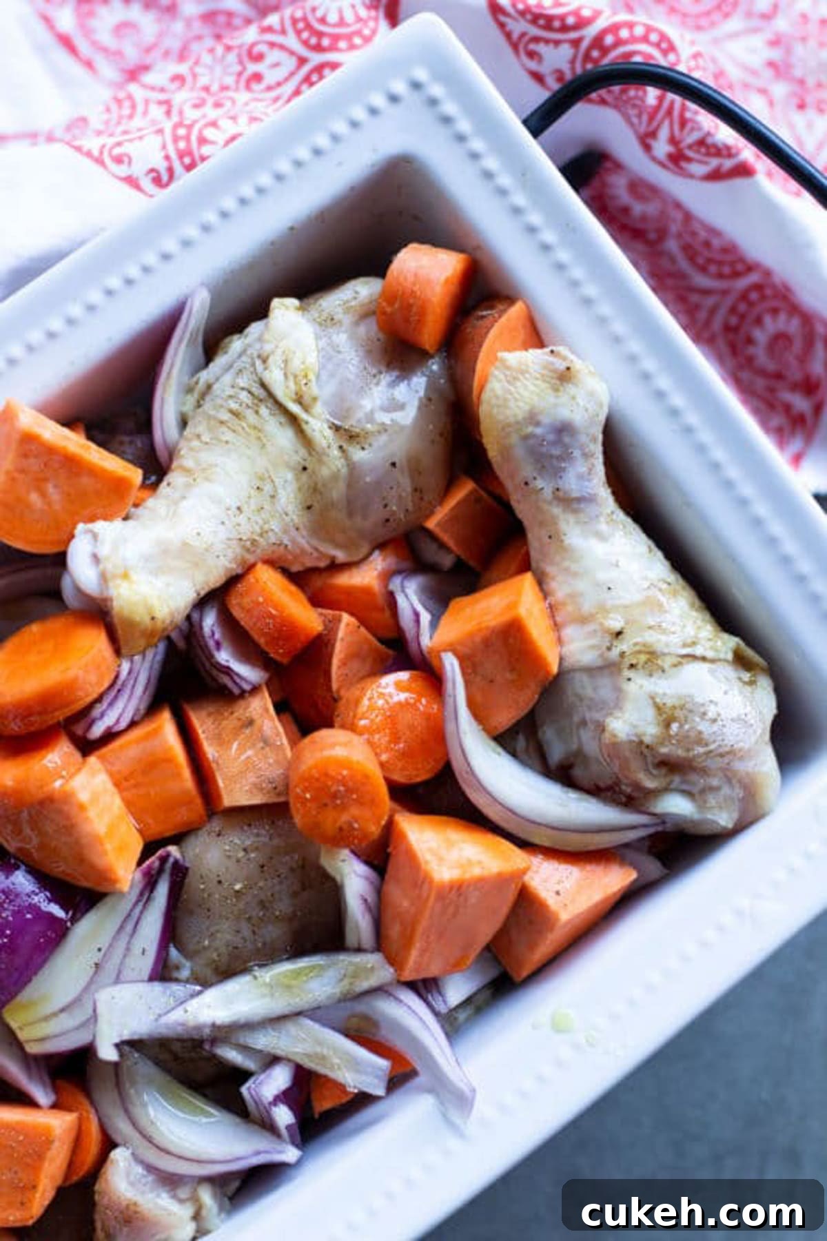 White dish containing seasoned drumsticks with sweet potatoes, carrots, and red onions drizzled with olive oil.
