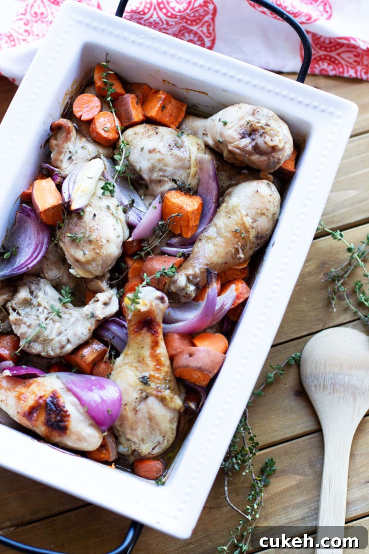 White dish containing roasted chicken drumsticks with sweet potatoes, carrots, and red onions topped with fresh thyme.