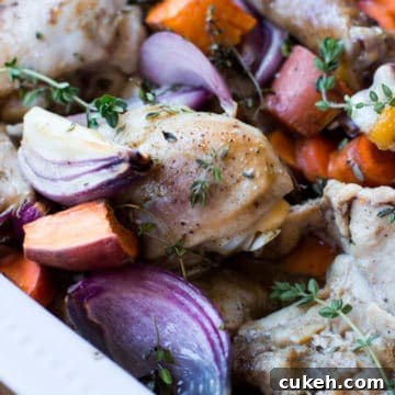 White dish containing roasted chicken with sweet potatoes, carrots, and red onions topped with fresh thyme.