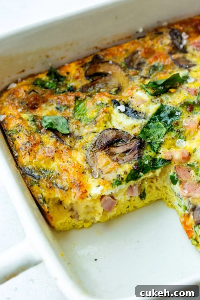 a baking dish of a baked egg, spinach and sauteed mushroom casserole with a serving missing so you can see the cross-section of the casserole