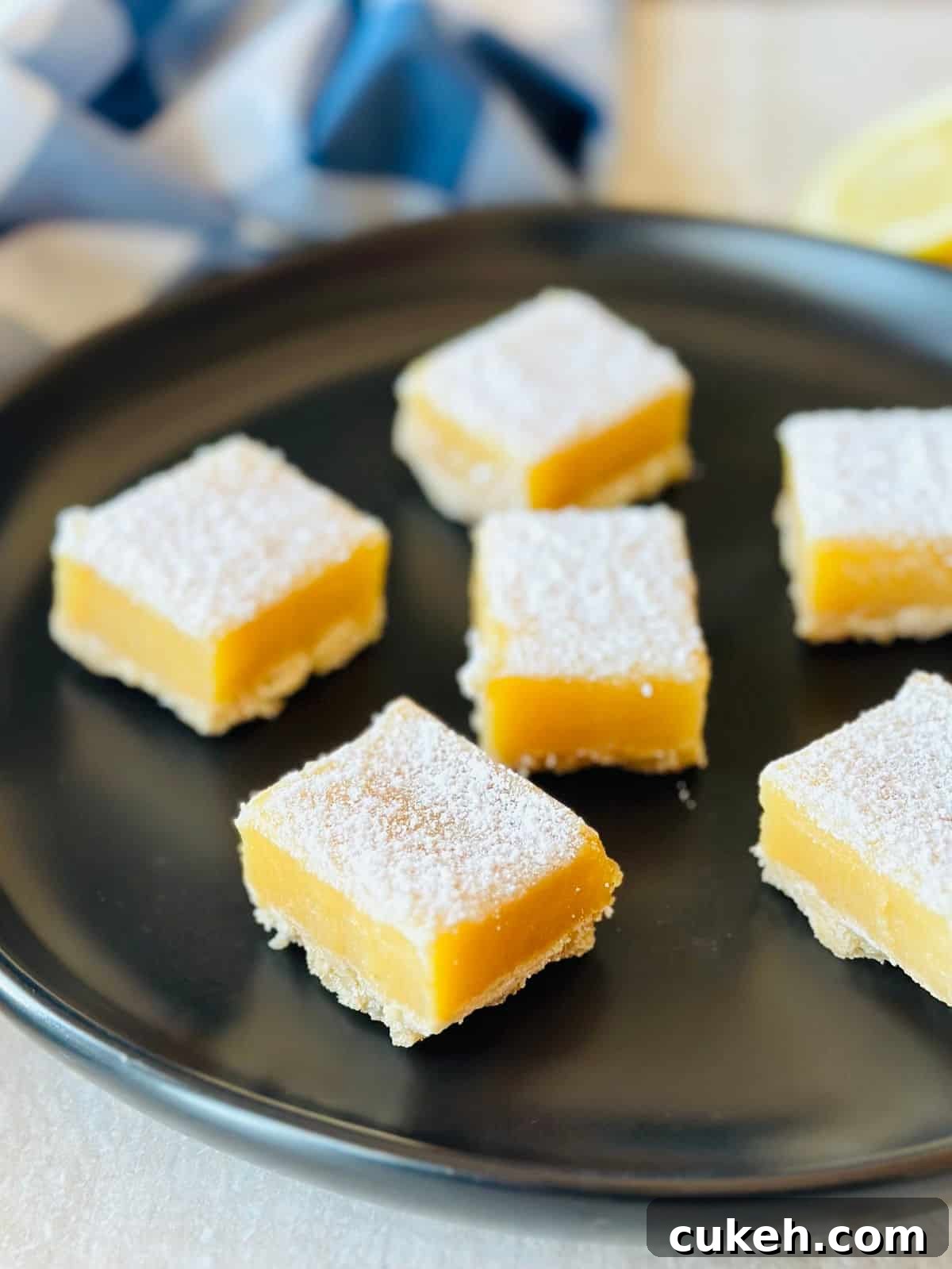lemon bars with graham cracker crust on black plate with sliced lemons and a blue napkin.