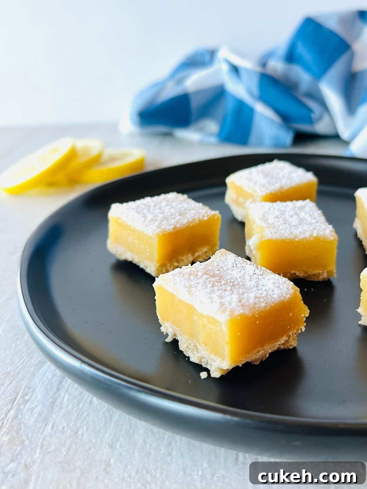 lemon cream cheese bars with graham cracker crust on black plate with sliced lemons and a blue napkin.