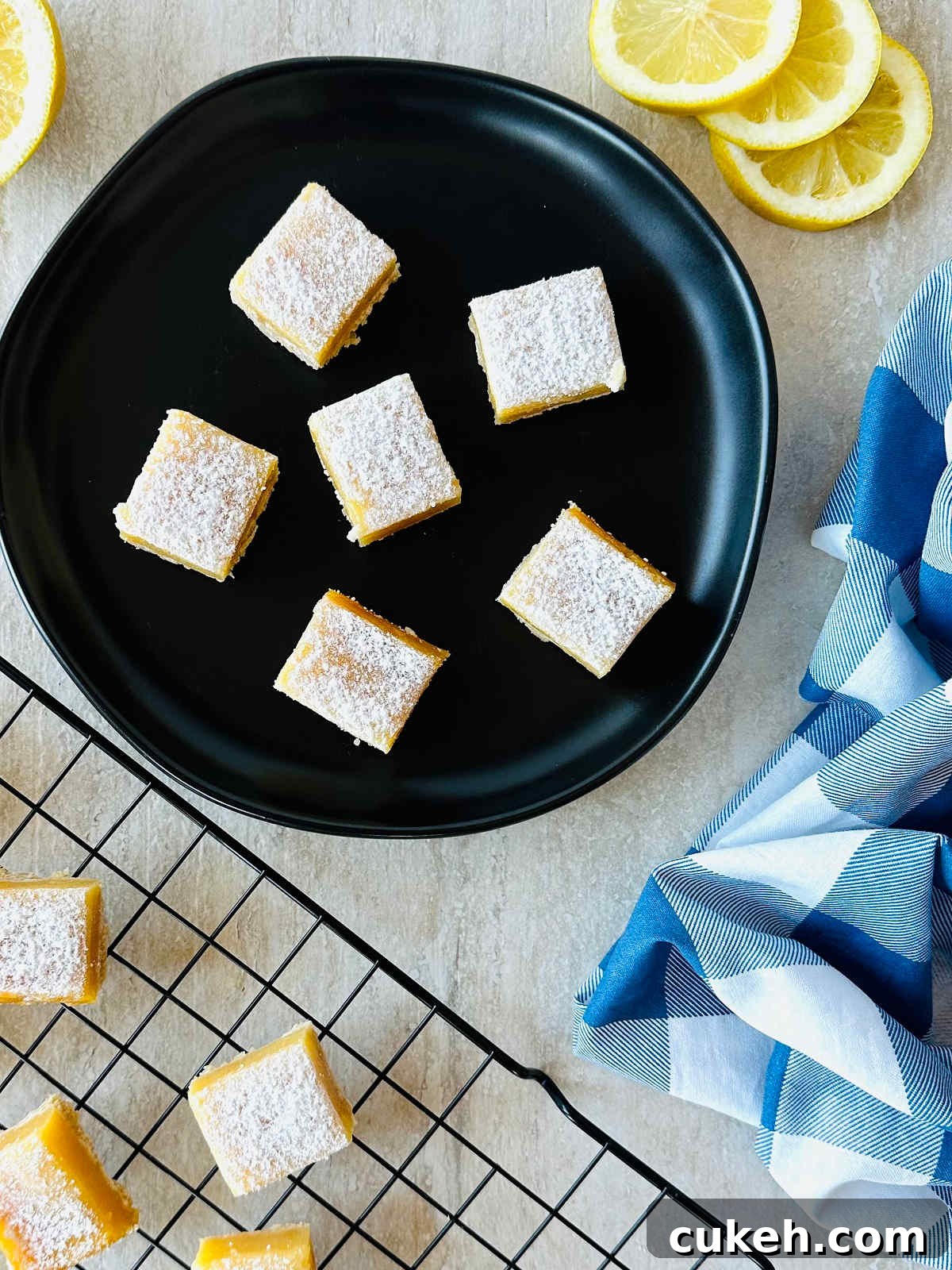 lemon bars with graham cracker crust on black plate with sliced lemons and a blue napkin.