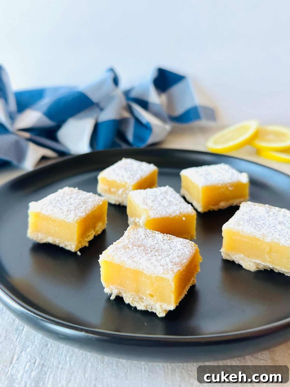 graham cracker lemon bars on black plate with sliced lemons and a blue napkin.