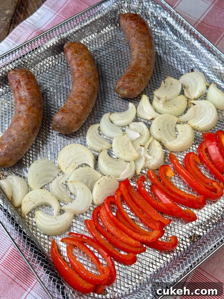 Three cooked Italian sausages in the air fryer basket in a row next to a row of cooked peppers and a row of onions.