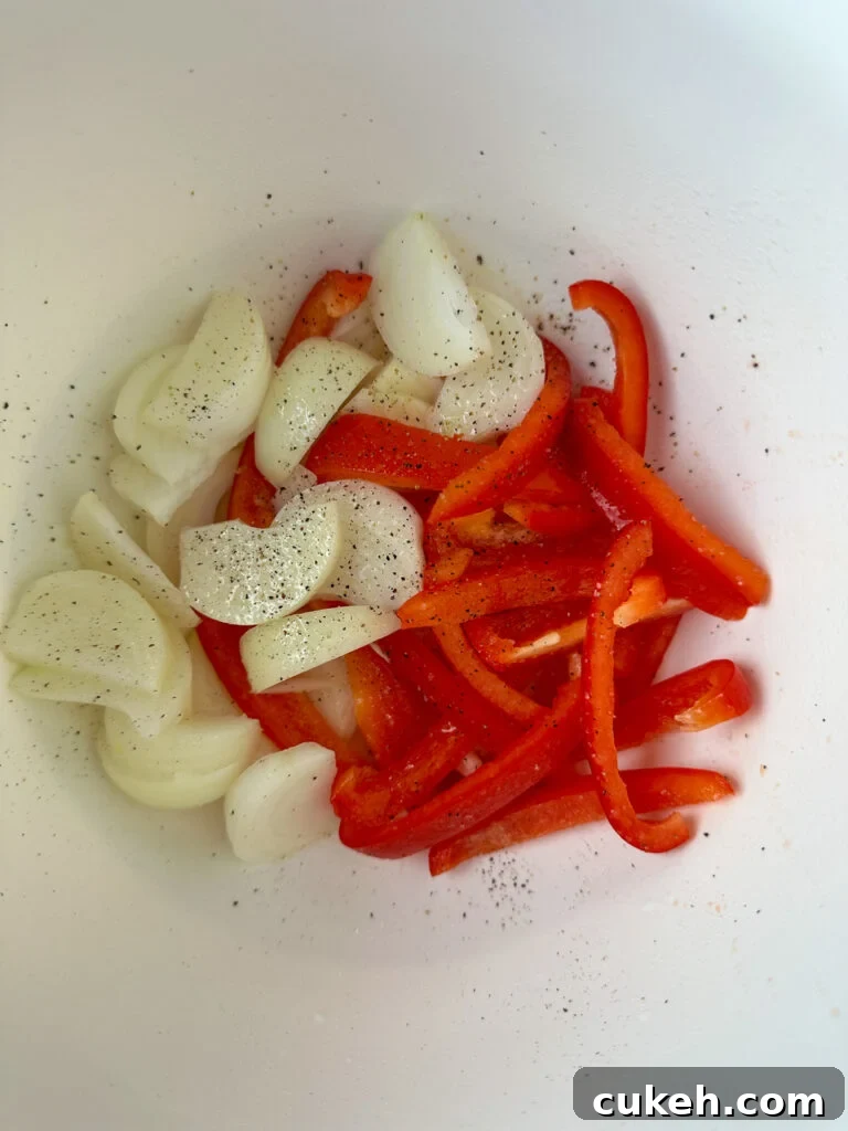 Slices of bell pepper and onion with a light coating of olive oil and salt and pepper sprinkled on top in a white mixing bowl.
