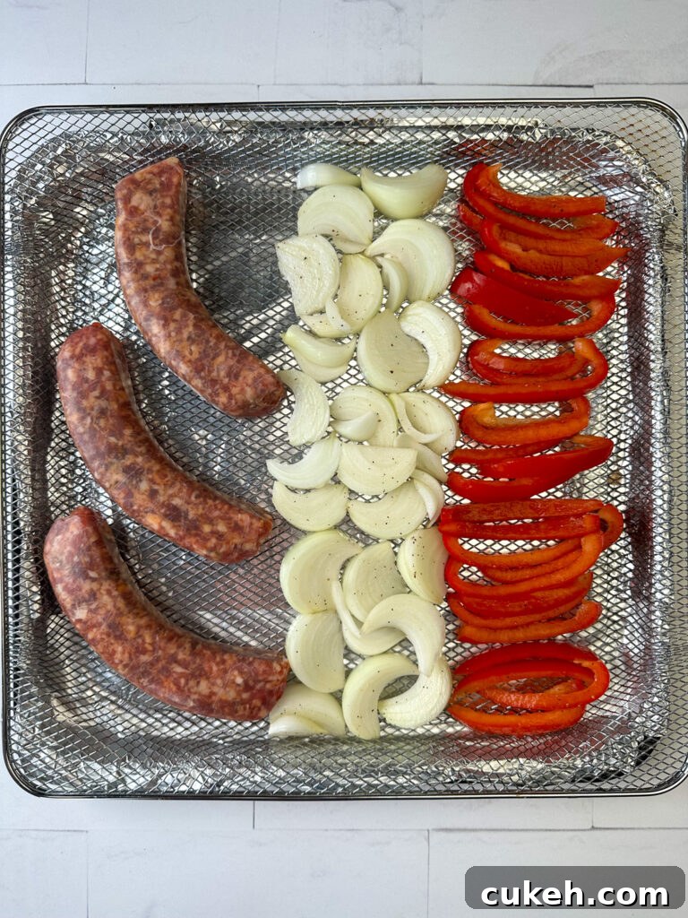 Three raw Italian sausages next to slices of red bell peppers in an air fryer basket on a white counter top.