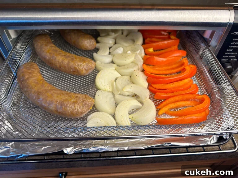Air fryer tray half out of the ninja foodi showing Italian sausages ready to flip in a row next to red peppers and onions with foil underneath.