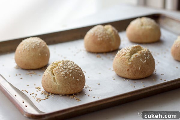 Fresh baked low carb sesame seed rolls with split tops on a parchment lined baking sheet placed on a white marble counter.