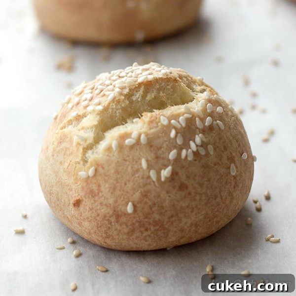 Round low carb roll with sesame seeds and a split top on a piece of parchment with another roll in the background.