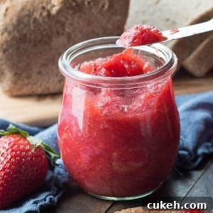 A knife with strawberry jelly on a jar of opened jelly in front of a blue napkin and a sliced loaf of bread on a cutting board.