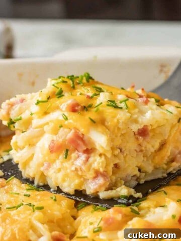 Slice of baked ham and cheese hash brown casserole on black spatula above white casserole dish.