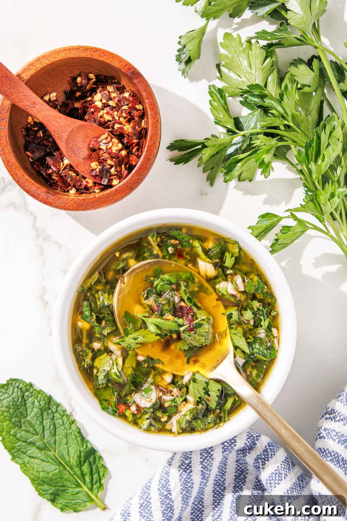 Fresh Mint Chimichurri Sauce Recipe for Grilled Meats & Veggies 2 A bowl of mint chimichurri with a wooden spoon, next to a small bowl of chili flakes and a striped napkin, on a white surface.