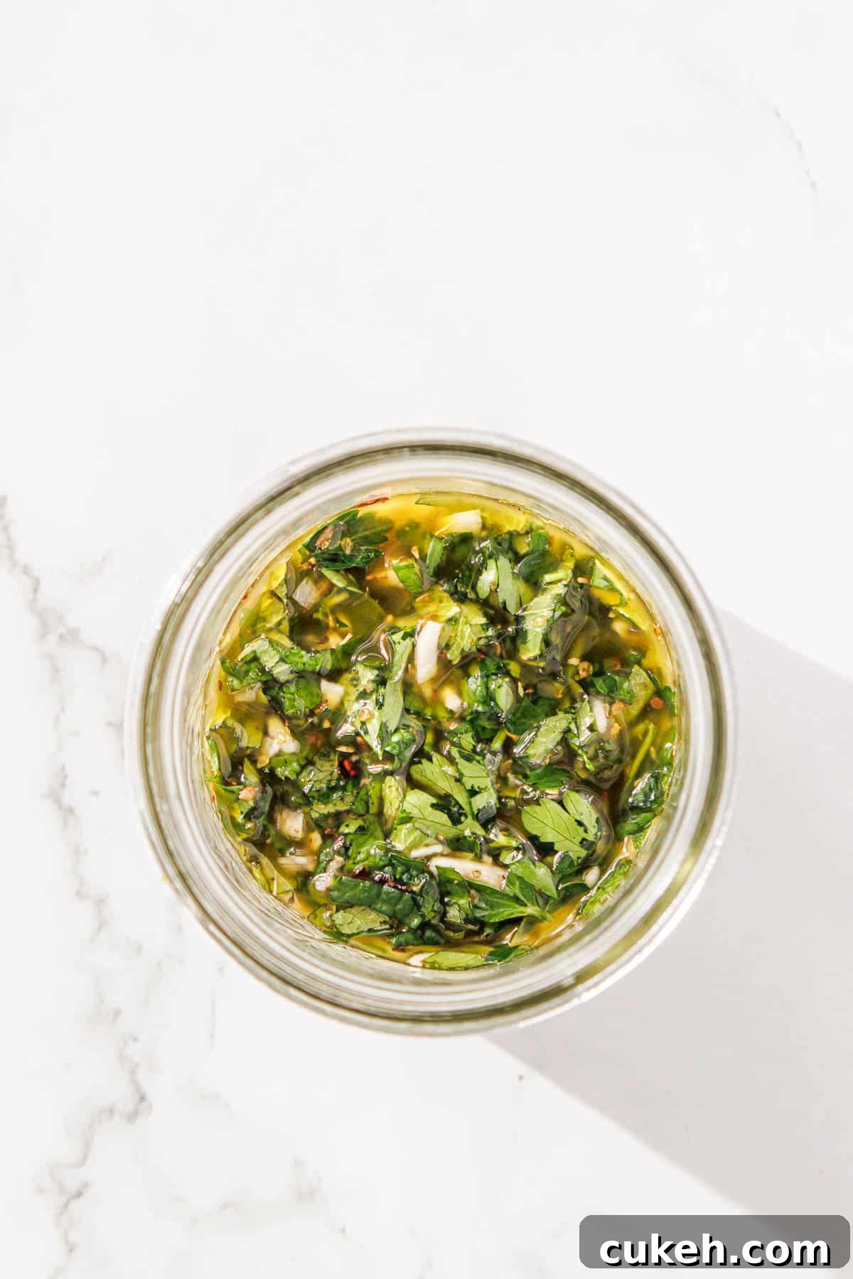 Fresh Mint Chimichurri Sauce Recipe for Grilled Meats & Veggies 6 Top view of a jar containing olive oil infused with herbs and spices, placed on a white marble surface.