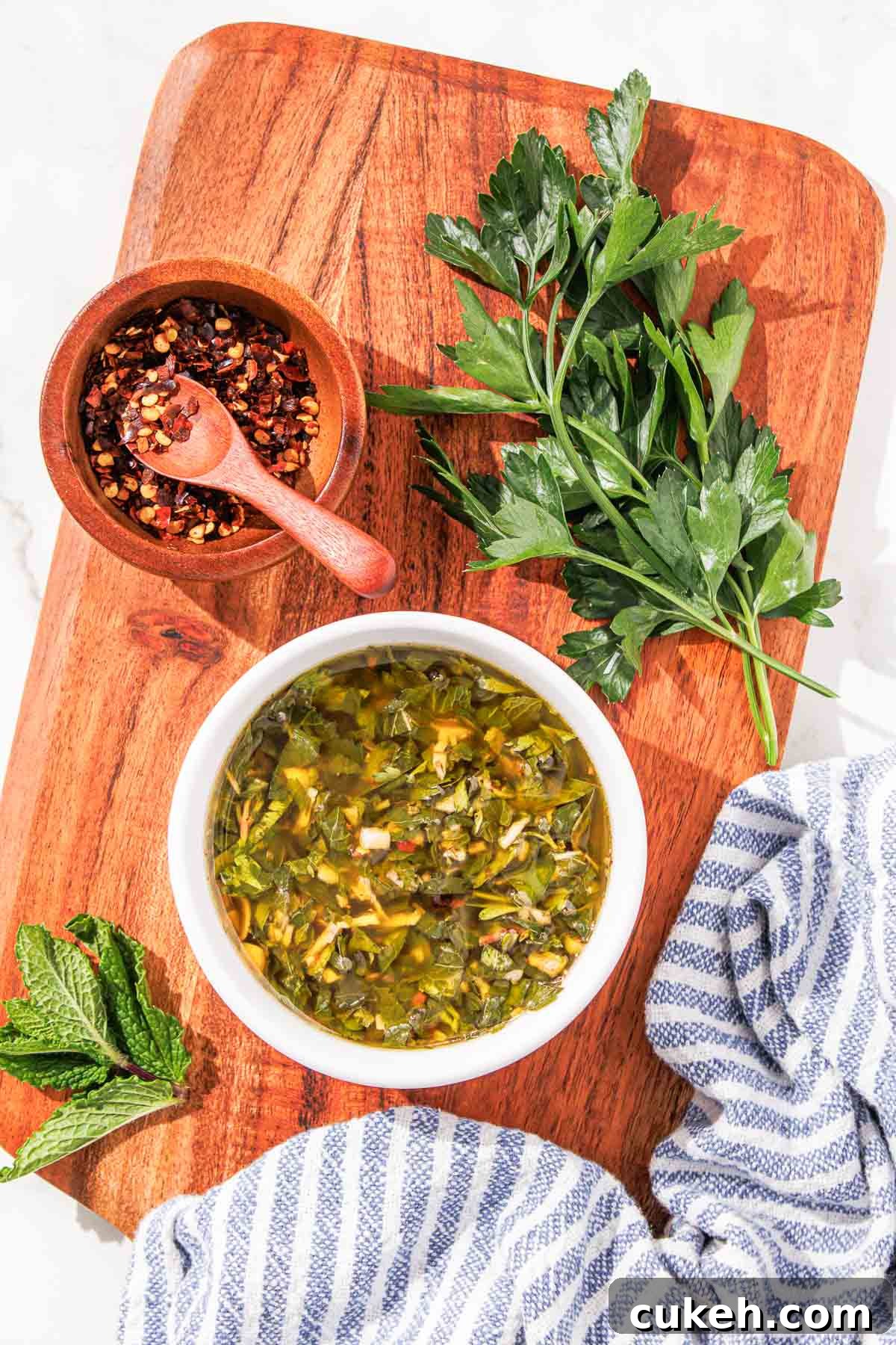 Fresh Mint Chimichurri Sauce Recipe for Grilled Meats & Veggies 8 A bowl of mint chimichurri sauce on a wooden cutting board, accompanied by fresh herbs and a bowl of spices. a striped cloth is partially visible.