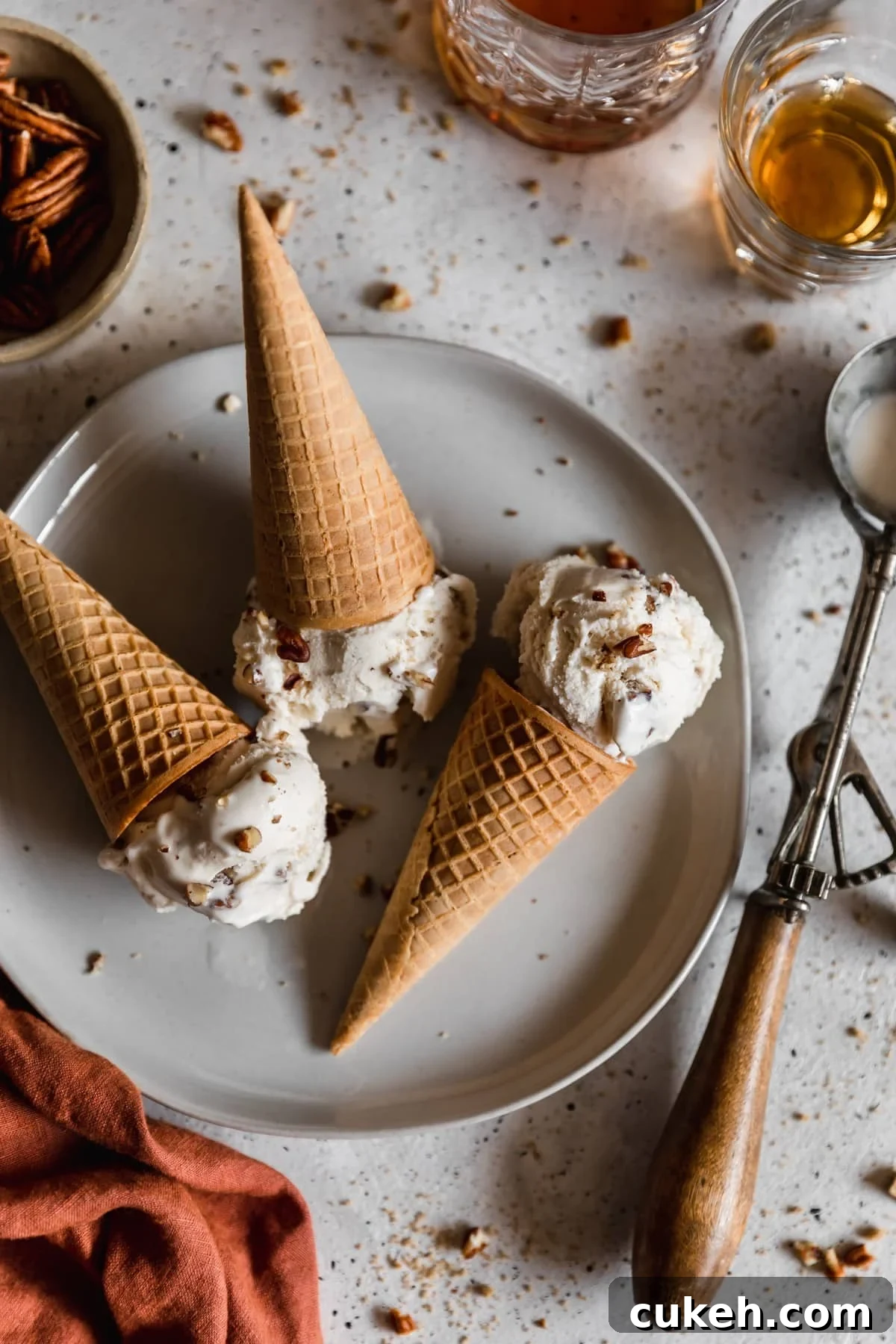 A closeup overhead image of three scoops of bourbon butter pecan ice cream with cones on a white plate next to a vintage ice cream scoop, two glasses of whiskey, and burnt orange linen. The plate is on a white speckled table dusted with chopped pecans.