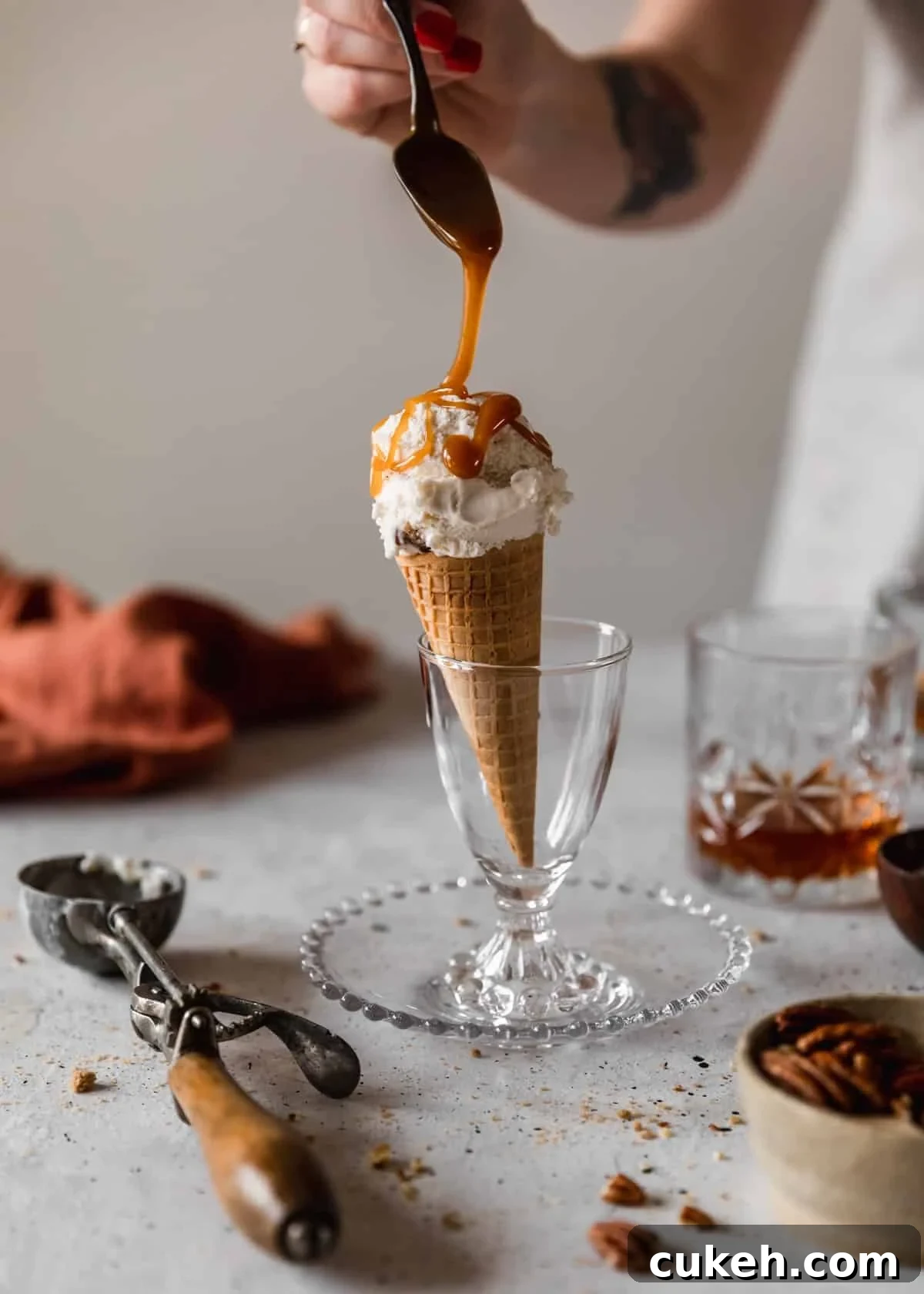 A side image of an ice cream cone in a glass cup on a white speckled table next to a glass of bourbon, cream colored bowl of pecans, vintage ice cream scoop, and burnt orange linen. A woman wearing a beige apron is drizzling caramel over the ice cream with a vintage spoon.