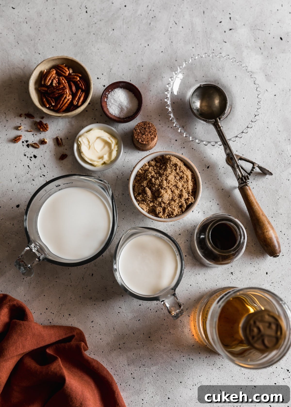 Two glass measuring cups with heavy cream and half-and half, a white bowl of brown sugar, a bottle of vanilla, a bottle of bourbon, and bowls of butter, salt, and pecans on a white speckled table. Next to the ingredients is a vintage ice cream scoop and dark orange linen.