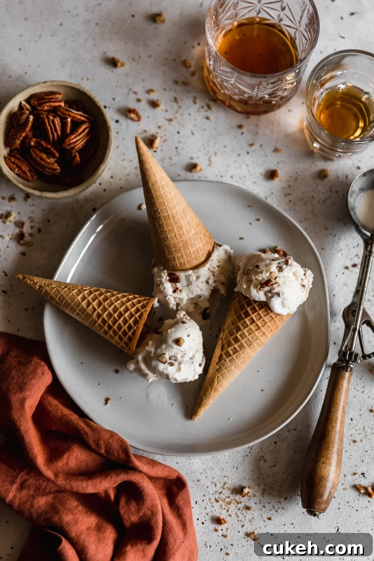 Three scoops of ice cream topped with cones on a white plate with a white and black speckled background. The plate is next to an old ice cream scoop, glass of bourbon, bowl of pecans, and orange linen.