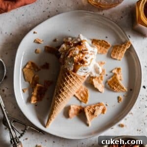 A closeup overhead image of a white plate topped with a butter pecan ice cream cone on a bed of ice cream cone shards with a white background.