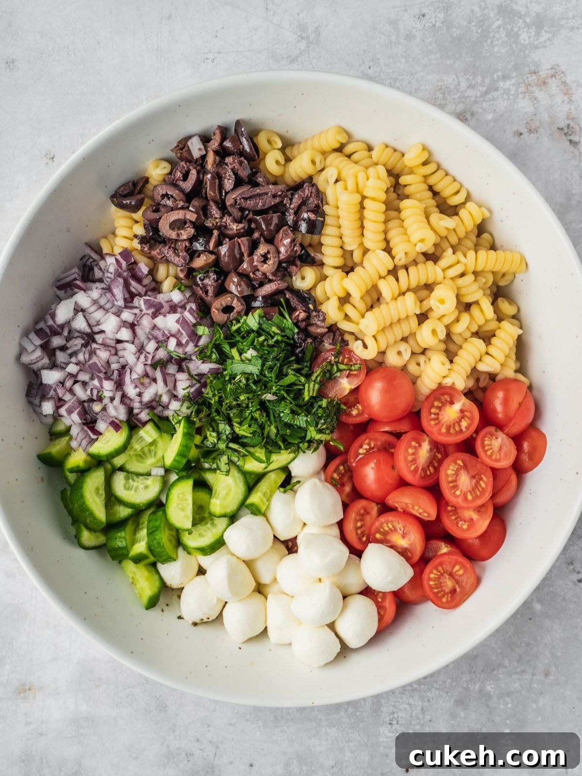 Recipe step showing a large serving bowl with cooked pasta, sliced cherry tomatoes, chopped cucumbers, diced onion, mozzarella balls and chopped herbs.