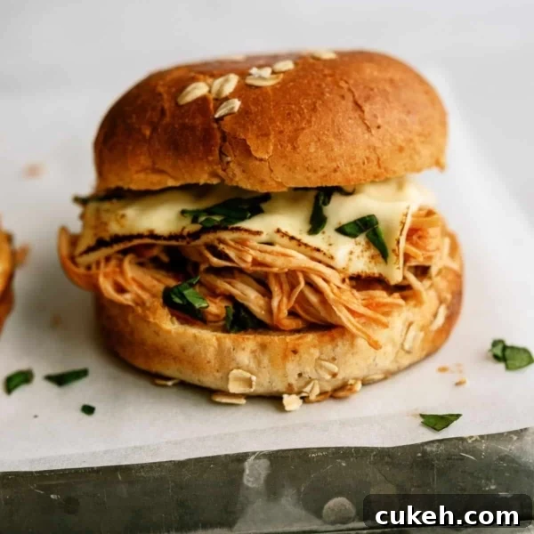 A sandwich with shredded chicken, melted cheese, and greens in a sesame seed bun, placed on white parchment paper.