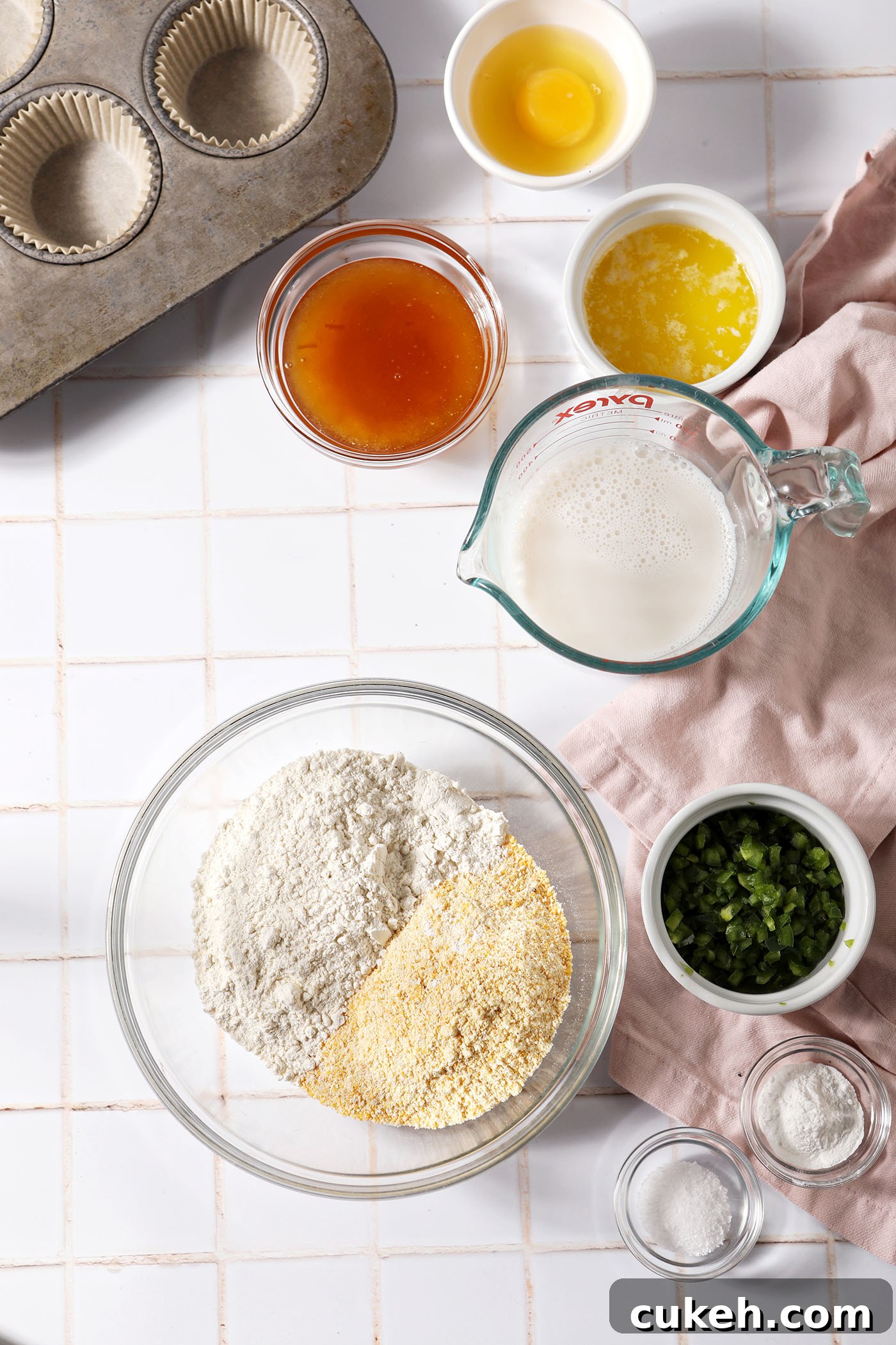 Honey Jalapeño Cornbread Muffins That Balance Sweet and Heat 3 Ingredients to make cornbread muffins with honey and jalapeño on a white tiled surface in bowls next to a pink linen