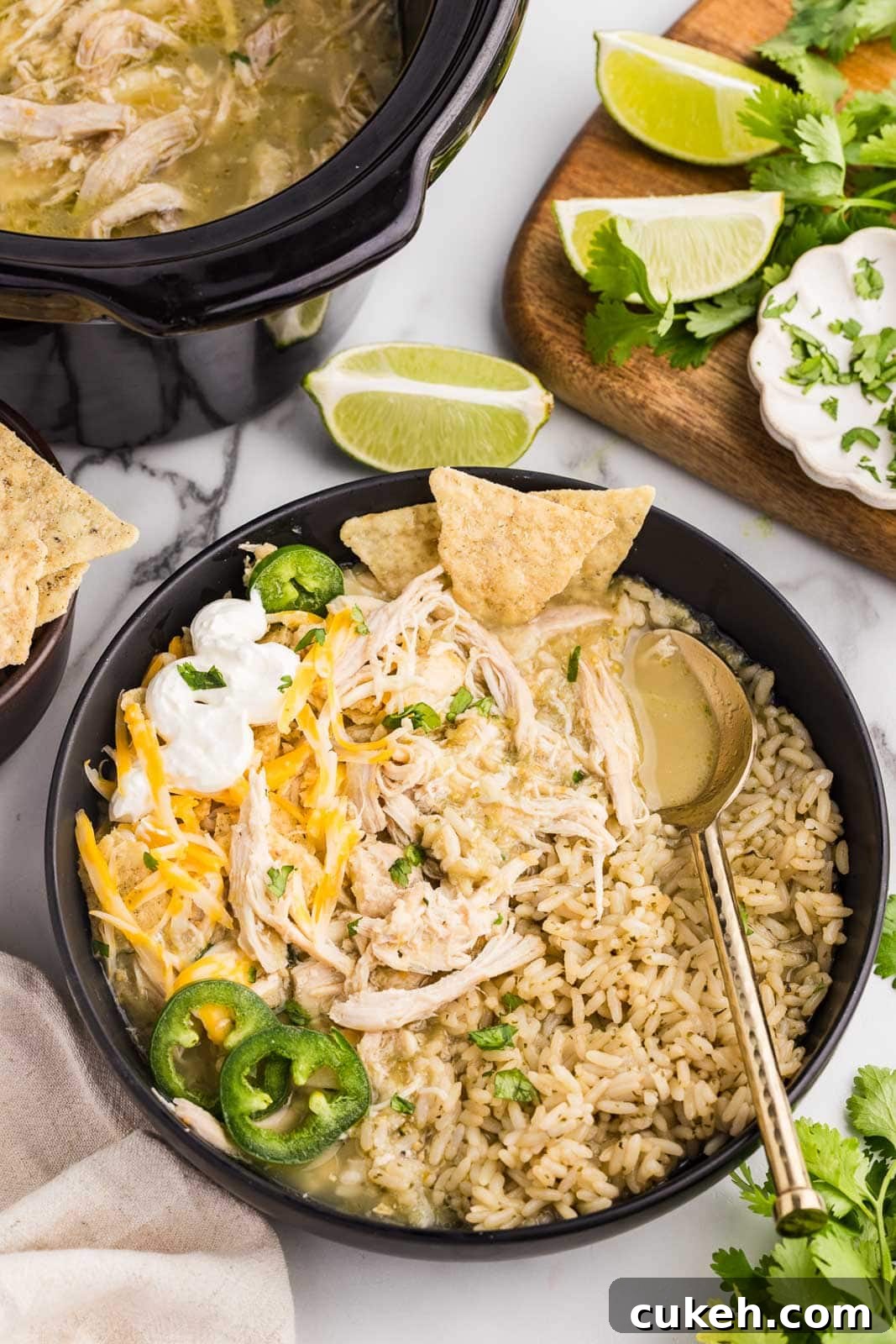 overhead view of black plate containing chile verde on rice with tortilla chips, jalapeno peppers cheese and sour cream, with limes and cilantro on a wooden cutting board with black slow cooker in background