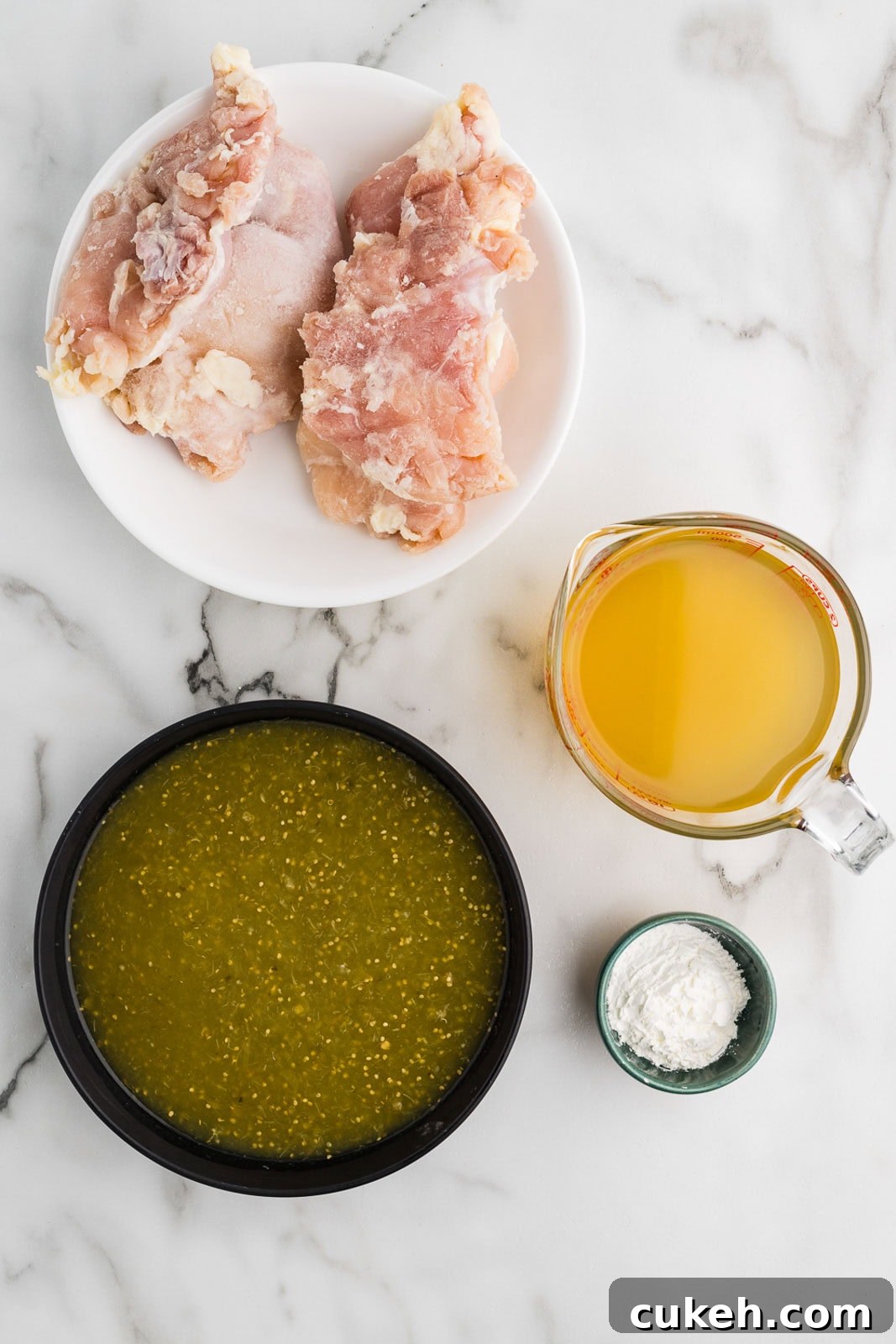 chile verde ingredients to include 2 chicken thighs on white plate, black bowl of salsa Verde, glass measuring cup of chicken broth and small bowl of cornstarch