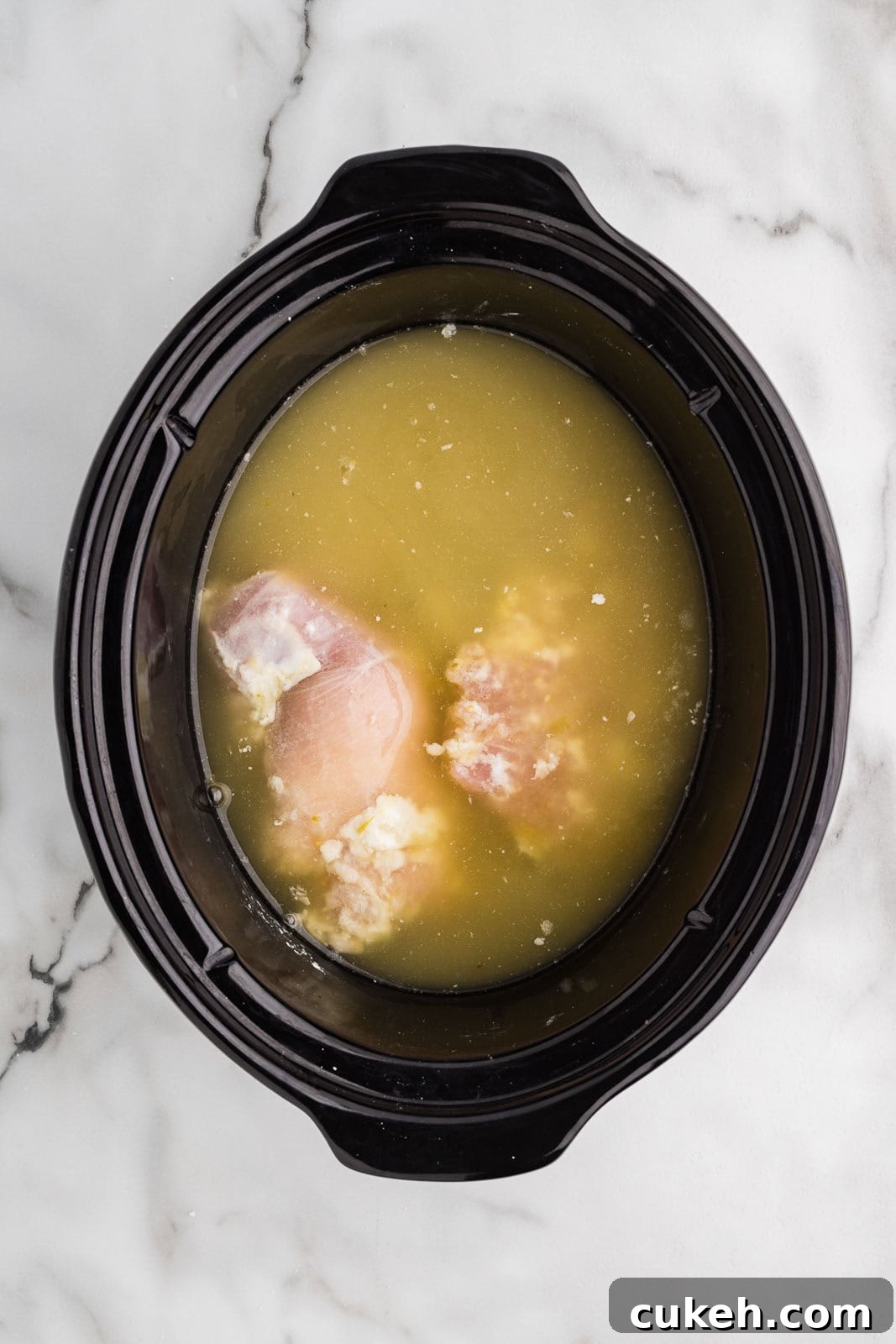overhead view of chicken thighs in black slow cooker covered with cornstarch, salsa and chicken broth