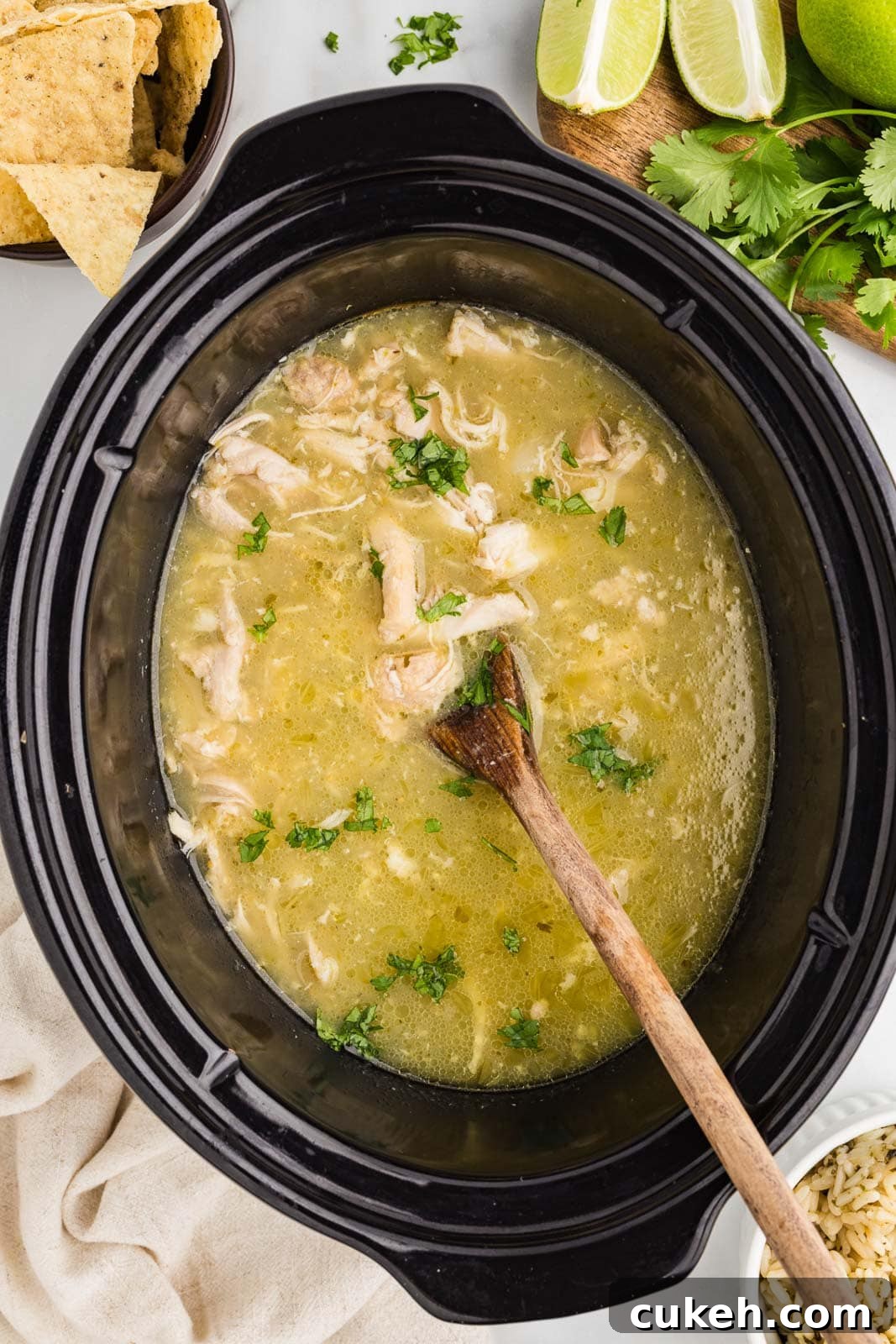 overhead view of chile verde in black slow cooker with wooden spoon inserted, surrounded by tortilla chips, limes and cilantro