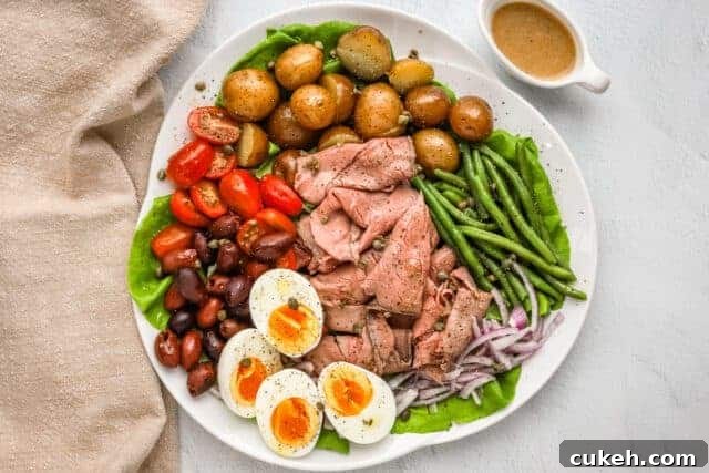 Beef nicoise ingredients are assembled on a platter with some capers, eggs, deli sliced roast beef, tomatoes, olives, potatoes, onions, green beans, and a Dijon mustard dressing.