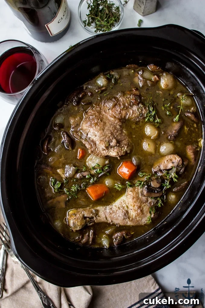 Overhead photo of Slow Cooker Coq Au Vin in a black slow cooker with a bottle of wine , glass of wine and salt and pepper shakers in background.