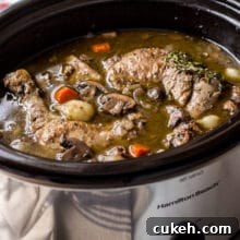 Coq Au Vin in a silver slow cooker with a bottle of wine and salt and pepper shakers in background.