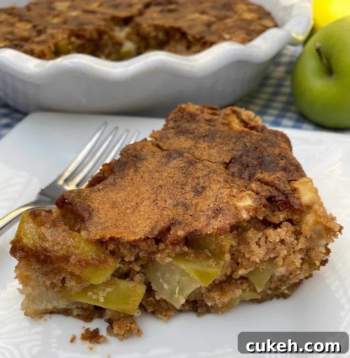 A wedge of apple cake with the baking dish and an apple in the background.
