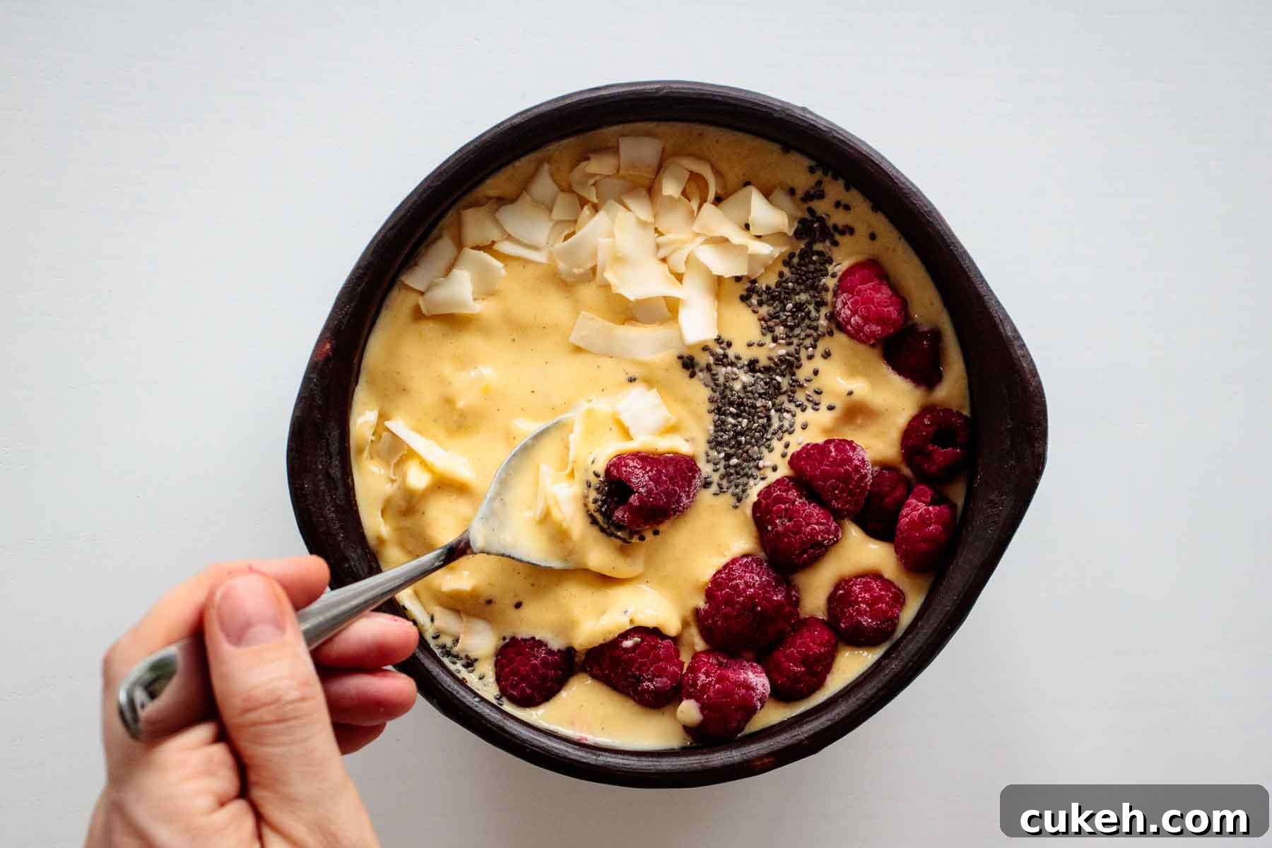 spoon scooping a breakfast bowl, with raspberry, chia and coconut flakes on top
