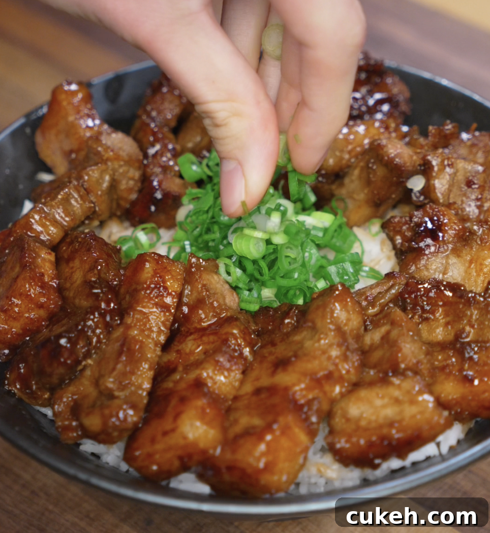 Sticky Glazed Pork Belly Bites: Sweet Savory Crispy Recipe 6 A hand sprinkles chopped scallions over a glossy bowl of sticky pork belly bites served on rice. The caramelized glaze and green onions add a vibrant finish to the rich dish.
