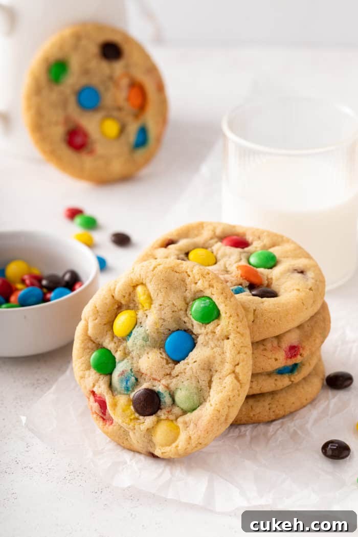 4 M&M cookies stacked on a piece of parchment paper in front of a glass of milk, with a 5th cookie leaning against the front of the stack.