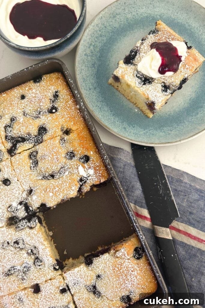 A photo of a tray of sourdough sheet pan pancakes with a piece missing from the tray. The piece of pancake is sitting on a blue plate to the right of the tray, garnished with vanilla yogurt and berry coulis.
