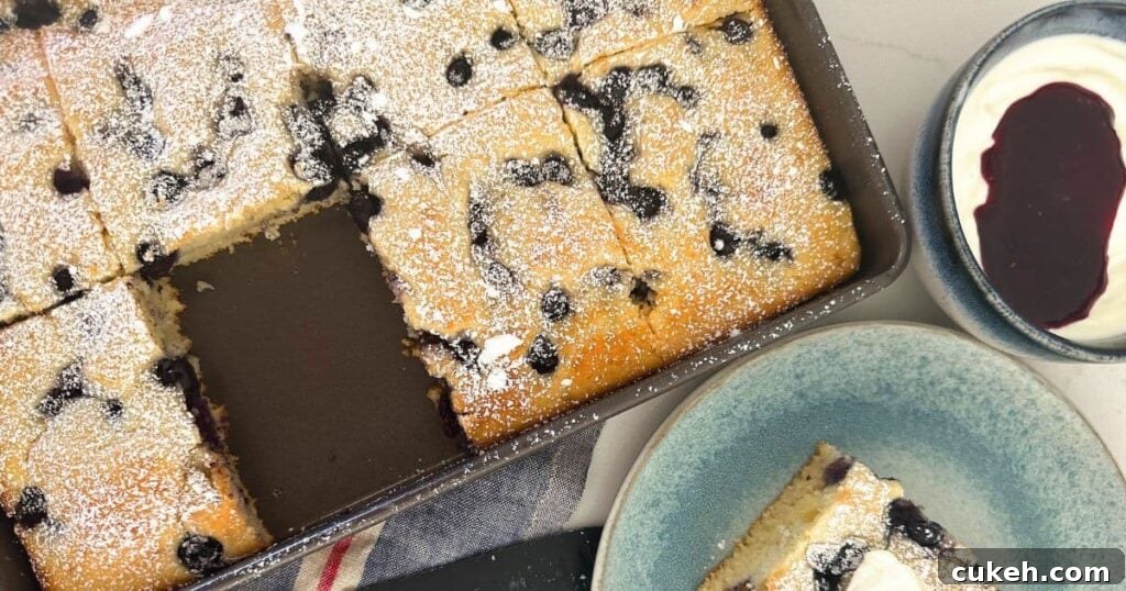 A photo of a tray of sourdough sheet pan pancakes with a piece missing from the tray. The piece of pancake is sitting on a blue plate to the right of the tray, garnished with vanilla yogurt and berry coulis.