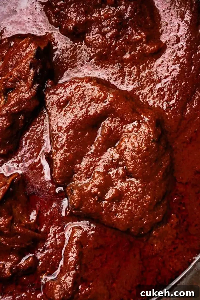 Close-up of tender beef birria simmering in a deep red chili-based sauce with visible oil and spices.