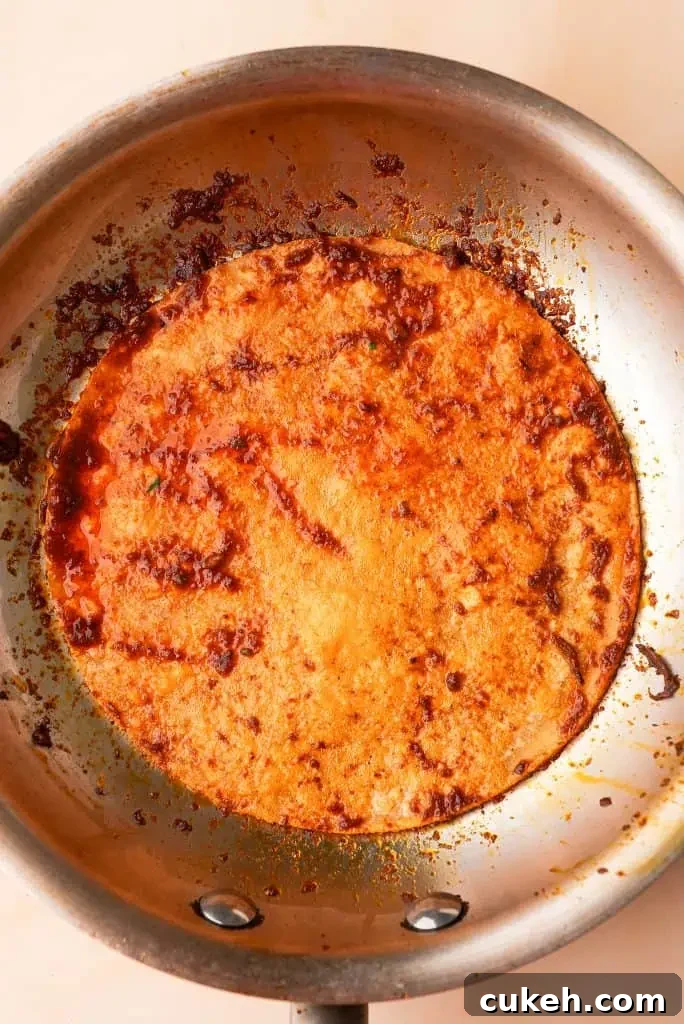 Corn tortilla dipped in birria consommé frying in a skillet with a rich red chile oil coating the surface.