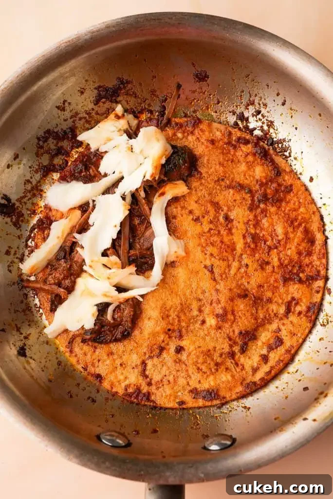 Corn tortilla in a skillet filled with shredded birria beef and melting cheese, partially folded as it cooks.