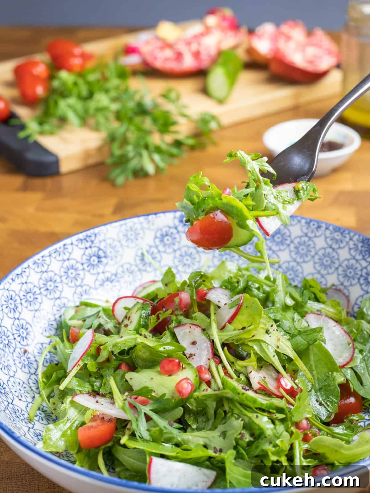 a blue and white decorated bowl filled with rocket salad and a chopping board with the ingredients in the background, all on a wooden table, with a forkful of salad being held up above the bowl.