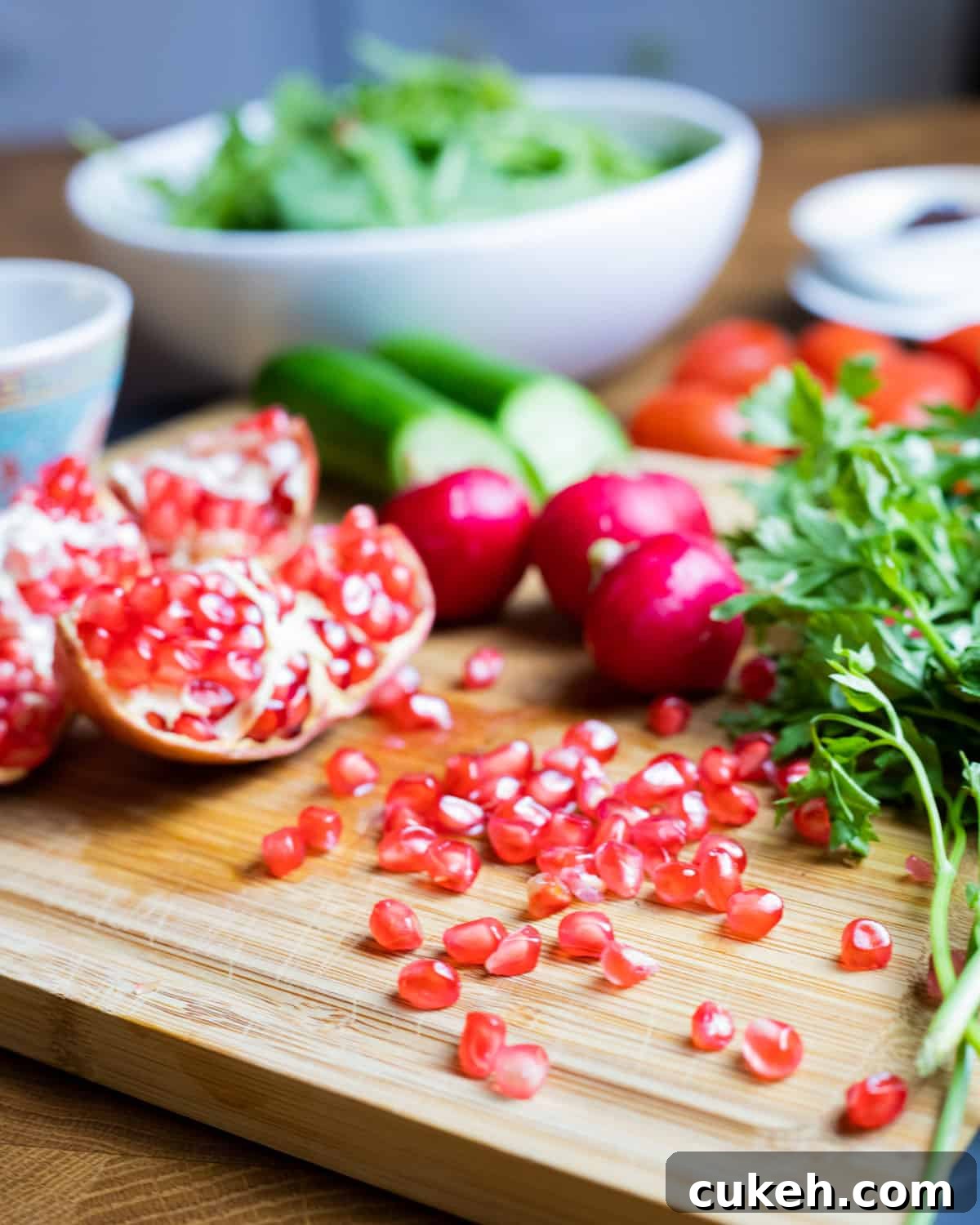 Photo of a chopping board with cut pomegranate and the seeds plus radishes, cucumbers, parsley and a bowl of rocket int he background.
