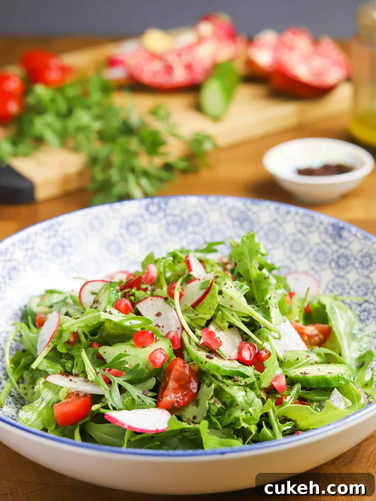 Photo of rocket salad in a pretty blue and white bowl on a wooden tabletop with a chopping board and veggies in the background.
