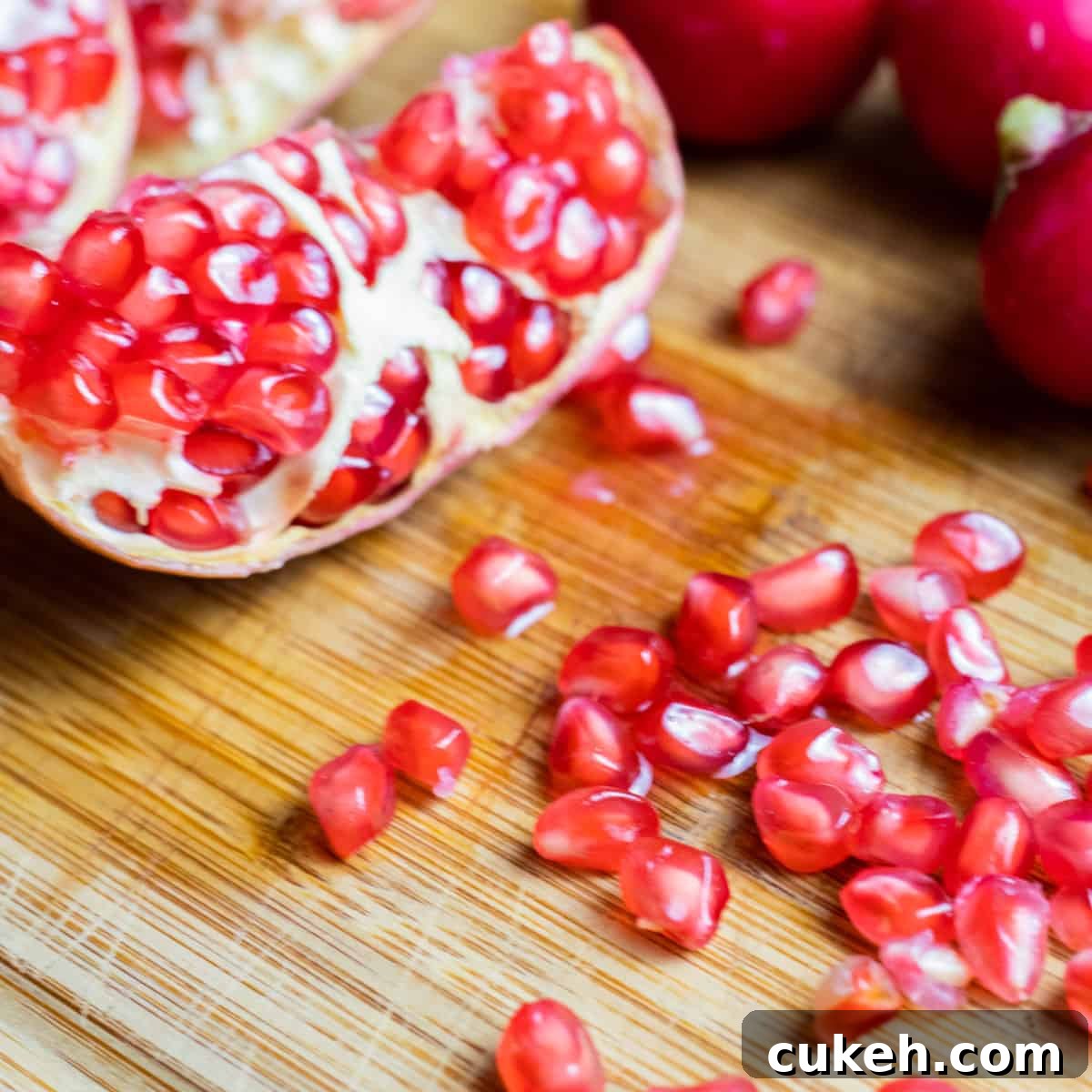 chopping board with a halved pomegranate and some pomegranate seeds scattered, with radishes and cucumber in the background.